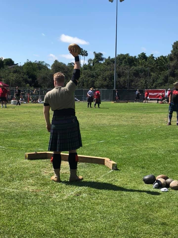 Image of D&aelig;rick Gr&ouml;ss, standing on a field at a highland games event, holding a stone over his head preparing to throw it.